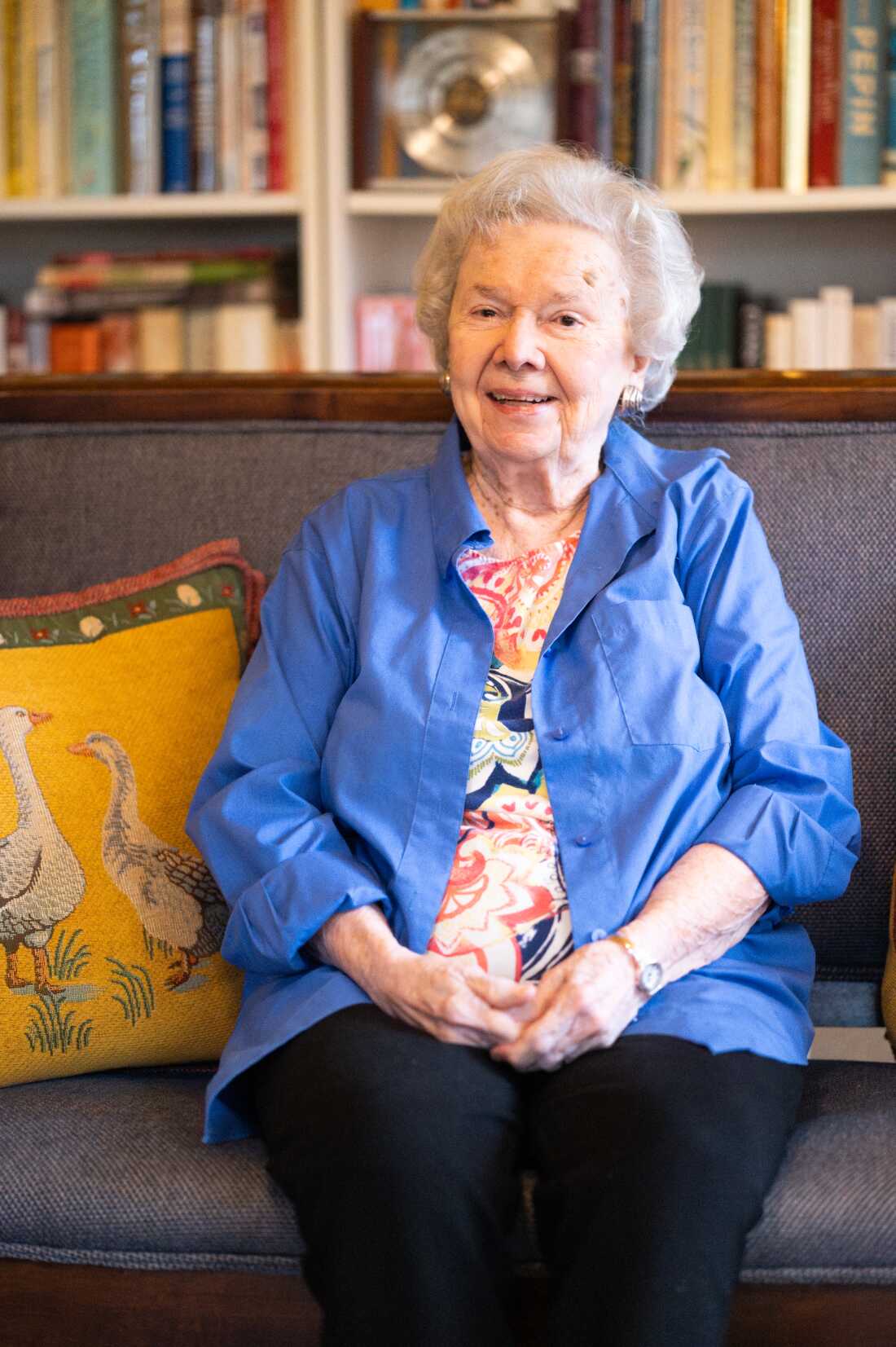 Barbara Briscoe, 93, sits on a vintage loveseat and book cases filled with books are seen behind her.