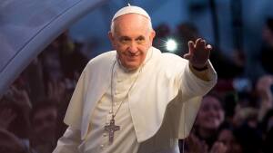 Pope Francis, wearing white, waves with his left hand with a crowd of people behind him.
