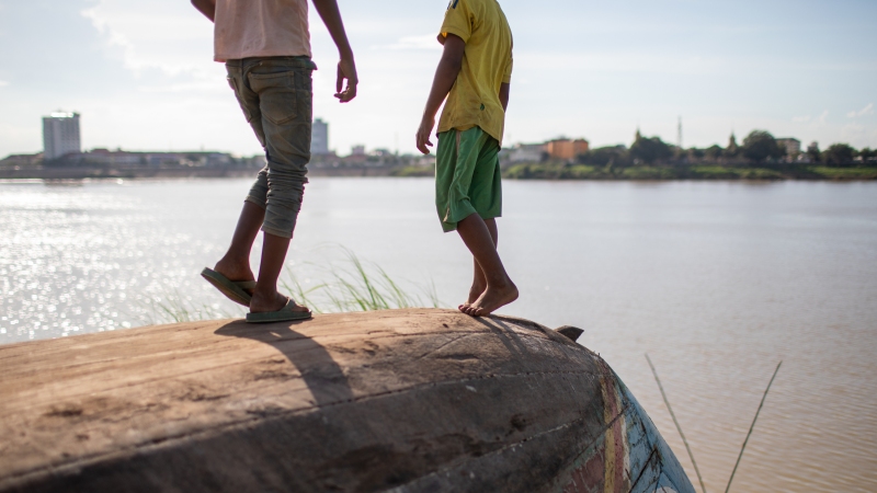 Two children stand on top of an overturned boat along a bank of the Mekong Delta.