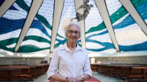 In this photo, Sister Rosita Milesi stands inside the Catedral Metropolitana de Brasilia, in the center of Brazil's capital. She's wearing a white, button-down shirt and glasses. Behind her is stained-glass artwork and an angel sculpture suspended from above.