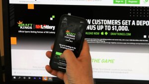 This photo shows a person's hand holding a smartphone with DraftKings on its screen. In the background is a computer monitor that is displaying DraftKings' website.