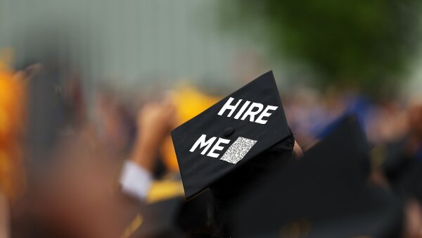 A black graduation cap with the words “Hire Me” on top of it. The background is a blurred view of graduates. 
