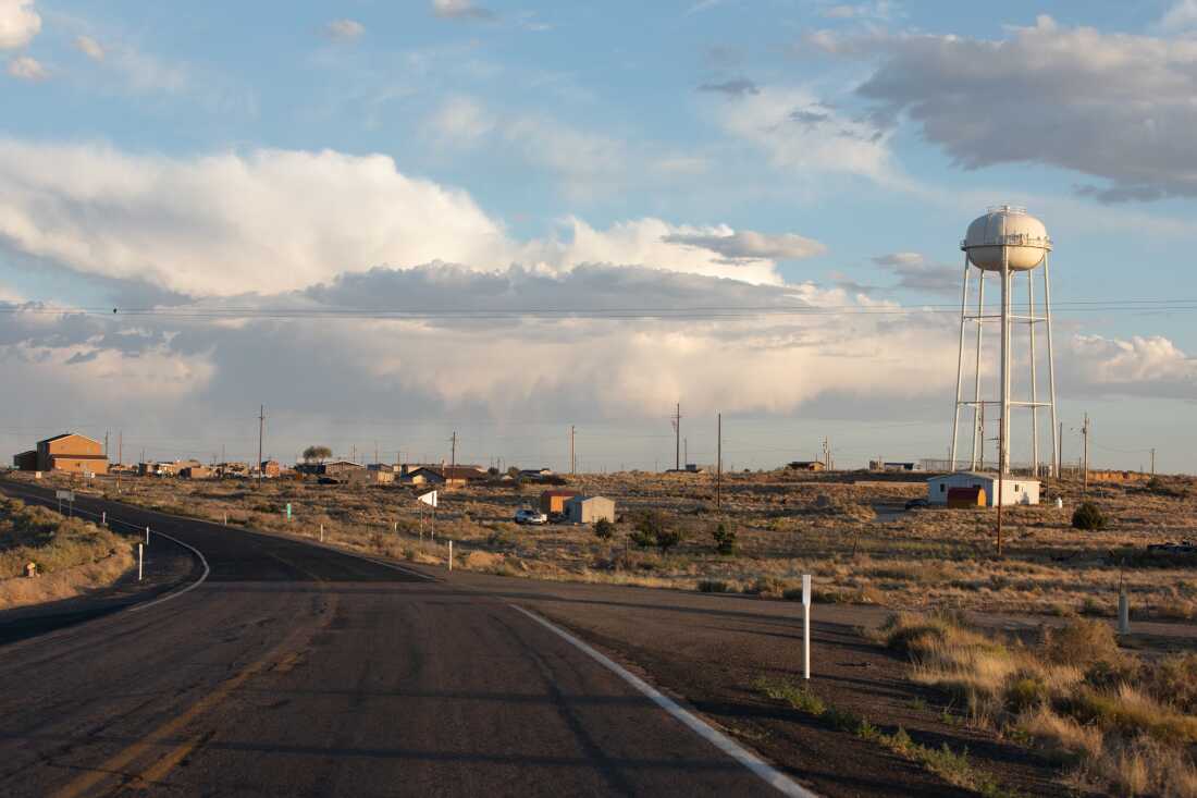 The late-afternoon sun shines over the village of Shongopovi. Buildings and homes are scattered across the dry landscape, and a road winds by on the left side of the frame. Wide white clouds float in the blue sky.