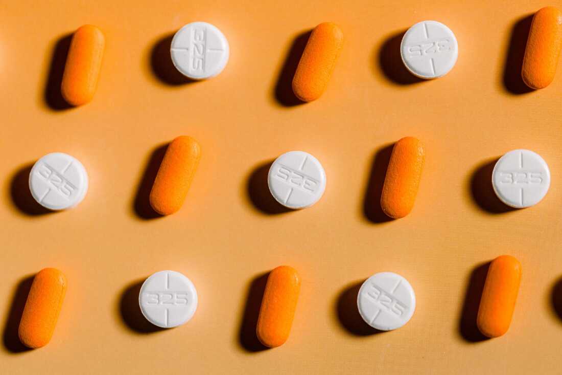 This close-up photo shows rows of pills alternating between acetaminophen (white circular pill) and ibuprofen (orange oval pill) on a bright orange background with a strong shadow.