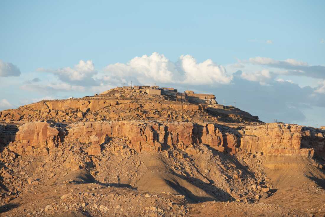 A village on the Hopi Reservation sits atop a mesa. The village is a cluster of short, boxlike brown structures situated at the top of a brown rock mesa.