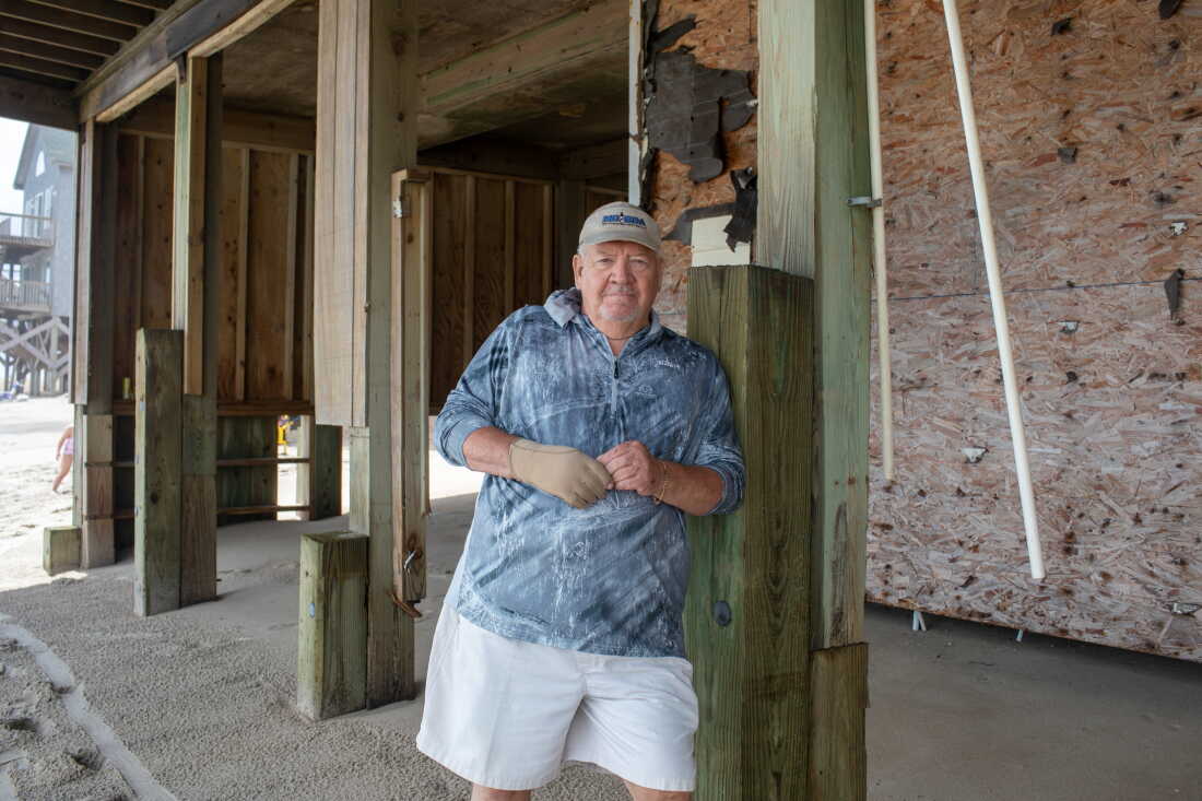 Bill King is standing by a house with exposed plywood and wooden beams. He's wearing white shorts, a gray shirt and a beige visor.