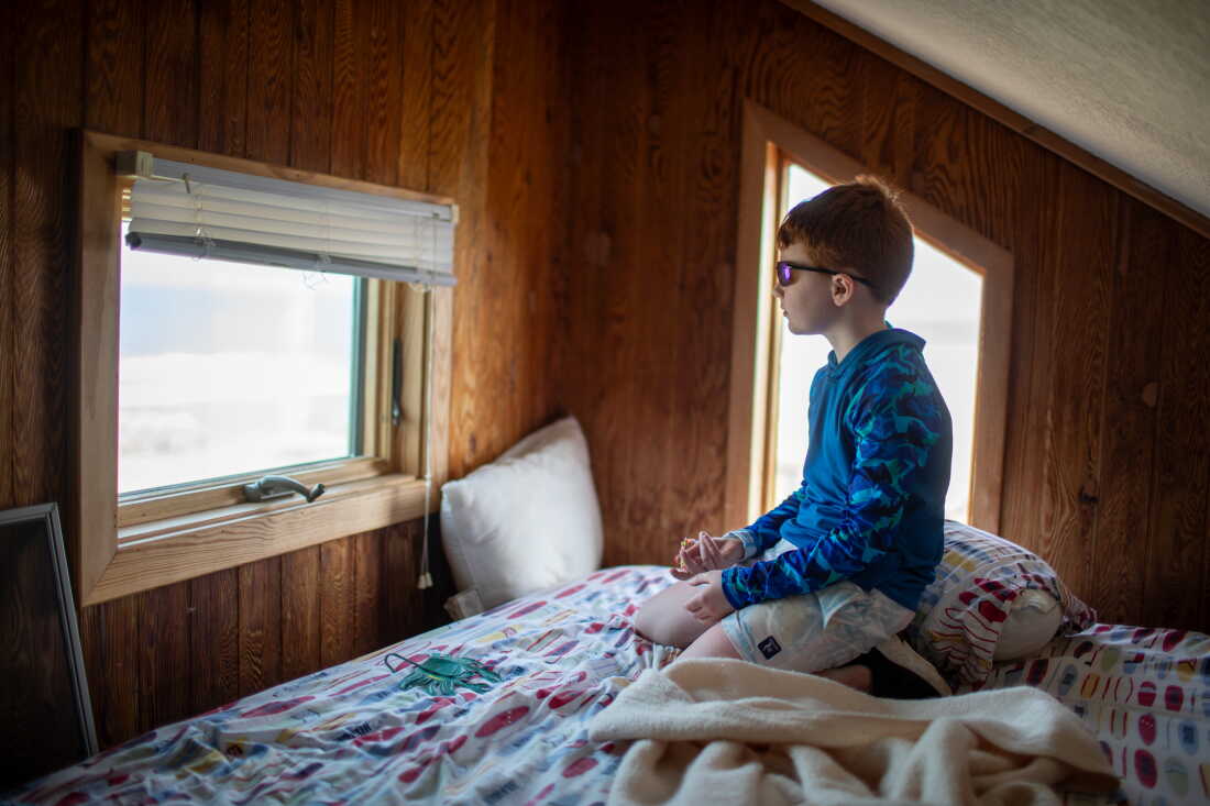 Clarke Lattimore, 8, sits on a bed as he looks out the window of his family's beach house in Buxton, North Carolina, in May. The boy is wearing sunglasses, a long-sleeved blue shirt and light-colored shorts. Vertical slats of wood cover the walls.