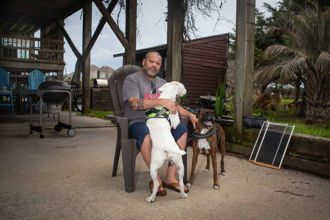 Brian Harris sits outdoors on a plastic chair. A white dog is climbing into his lap, and Harris is touching the back of a brown dog that's standing beside the chair.