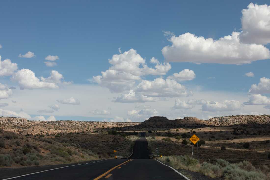 Fluffy white clouds hang in a blue sky above a lonely hillside road that winds through the Hopi Reservation.