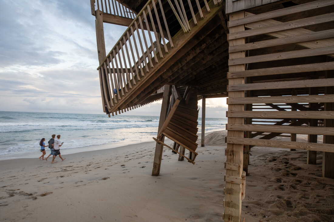 A damaged wooden deck-like porch hangs precariously over the sand on the Cape Hatteras National Seashore. The porch's wooden staircase is twisted and dangles over the sand. The wooden floor of the deck-like porch is tilted downward above the sand.