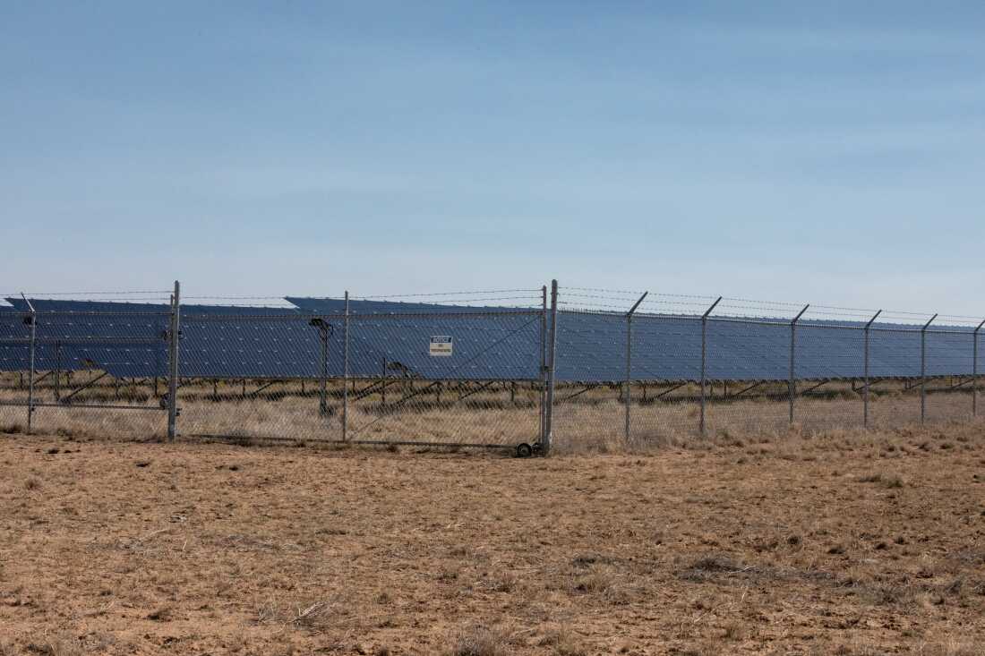Rows of solar panels stand behind a chain link fence and against a bright sky.