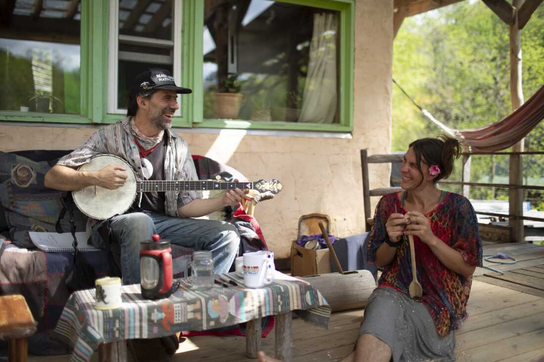 Zev Friedman and Chloe Vieira relax on their porch. Friedman plays a guitar while Vieira whittles a wooden spoon.