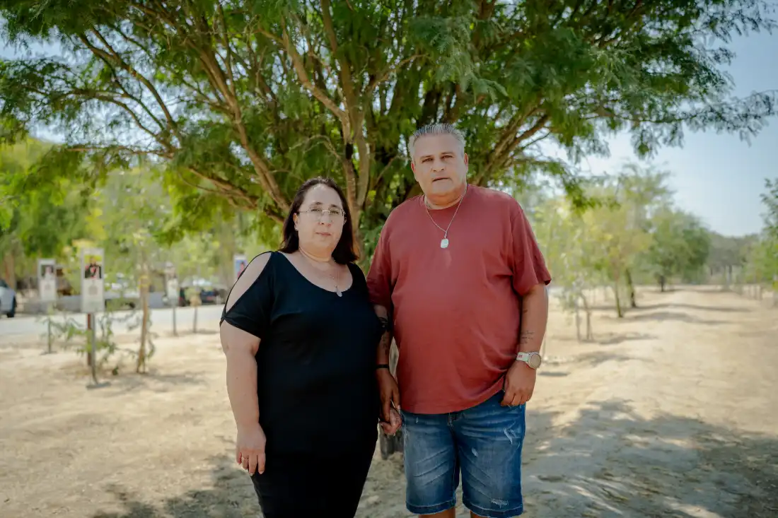 Meir and Kati Zohar hold hands as they pose for a portrait beneath a tree at the Nova music festival memorial site near Kibbutz Re'im, southern Israel, on Sept. 24, 2026. Their daughter, Bar Zohar, was killed during the Hamas-led assault on the festival on Oct. 7, 2023. (Erik Marmor for NPR)