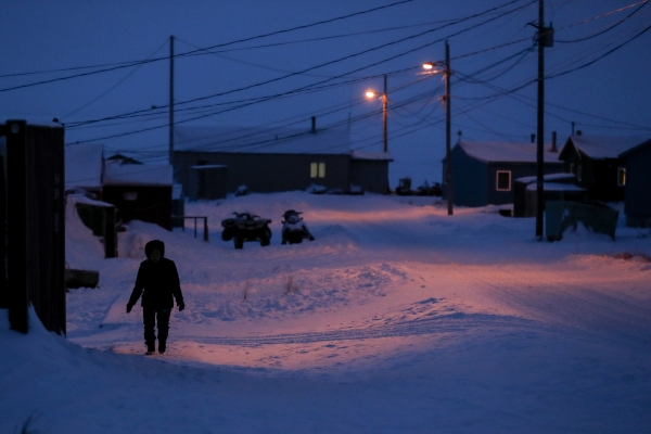 A woman dressed in winter clothing walks before dawn in Toksook Bay, Alaska, in 2020. The ground and the roofs of one-story buildings are covered in snow. The sky is dark, and electric lines run above the buildings.