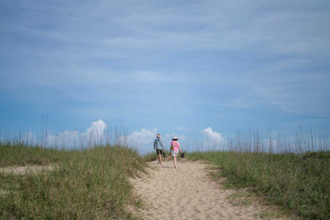 A man and a woman, their backs to the camera, walk barefoot along a sandy path at the coast. Tall green grass lines both sides of the path.