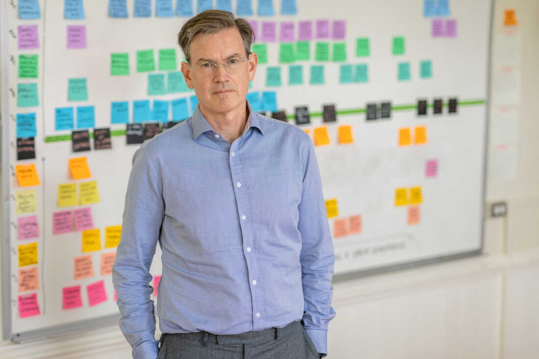 Mark Somerville stands in front of a whiteboard that's covered in rows of sticky notes, with each row a different color. Each note lists a task that the new college needs to complete before it opens.