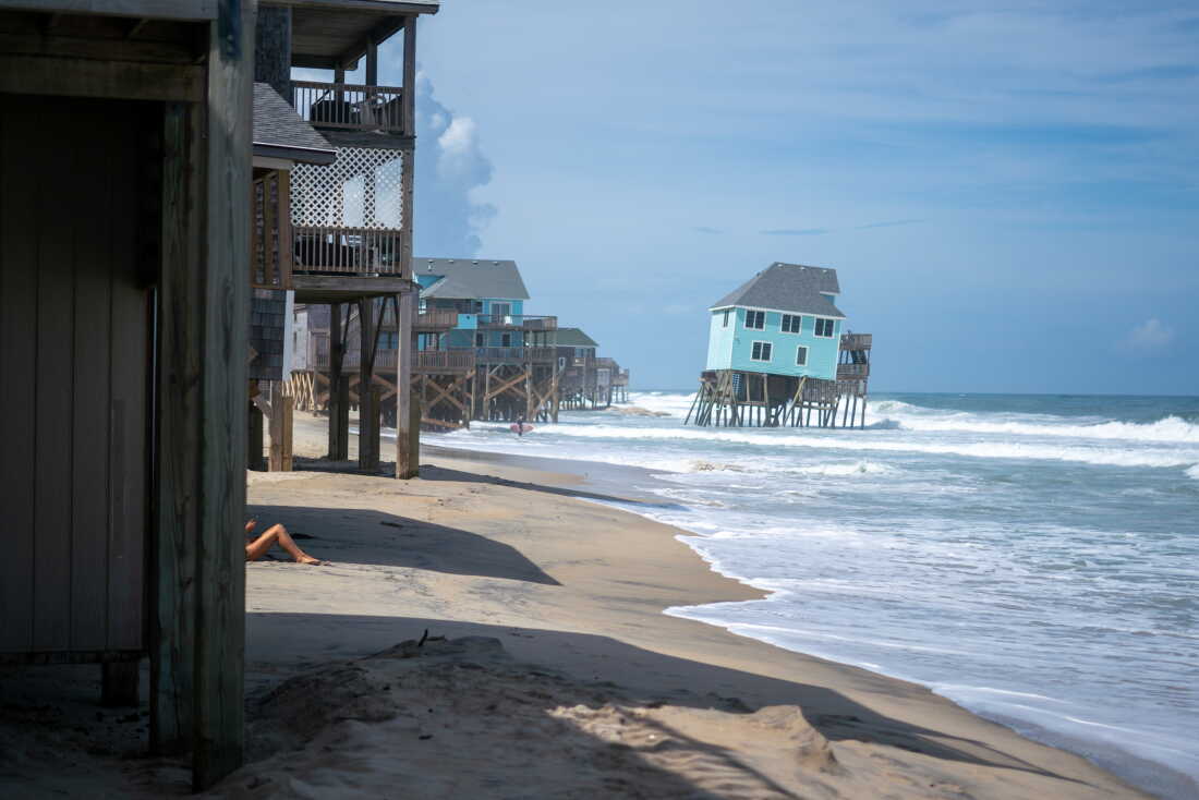 Multiple houses stand on wooden stilts on the beach along the coast. Water laps underneath the houses, at the bottom of the stilts. One house on stilts is leaning precariously toward the ocean.
