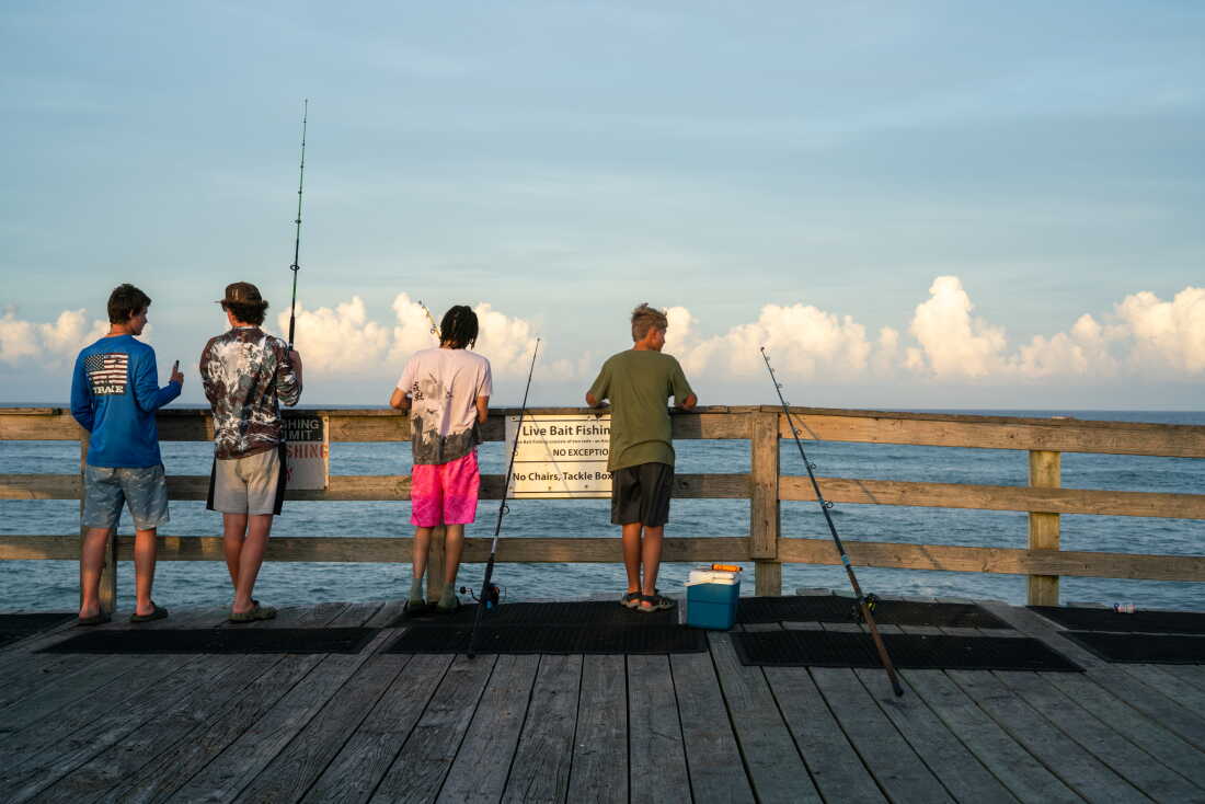 Four people, their backs to the camera, stand behind the wooden railing of a pier in Avon, North Carolina. Facing the water, they are fishing with fishing poles.