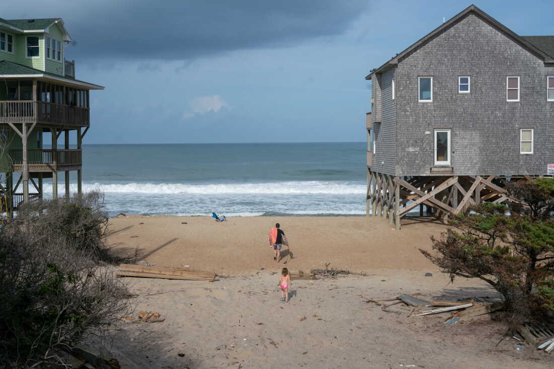 A man and girl clad in swimwear, their backs to the camera, walk cross the beach, toward the ocean. They are walking between two large multilevel houses that are perched on wooden stilts.