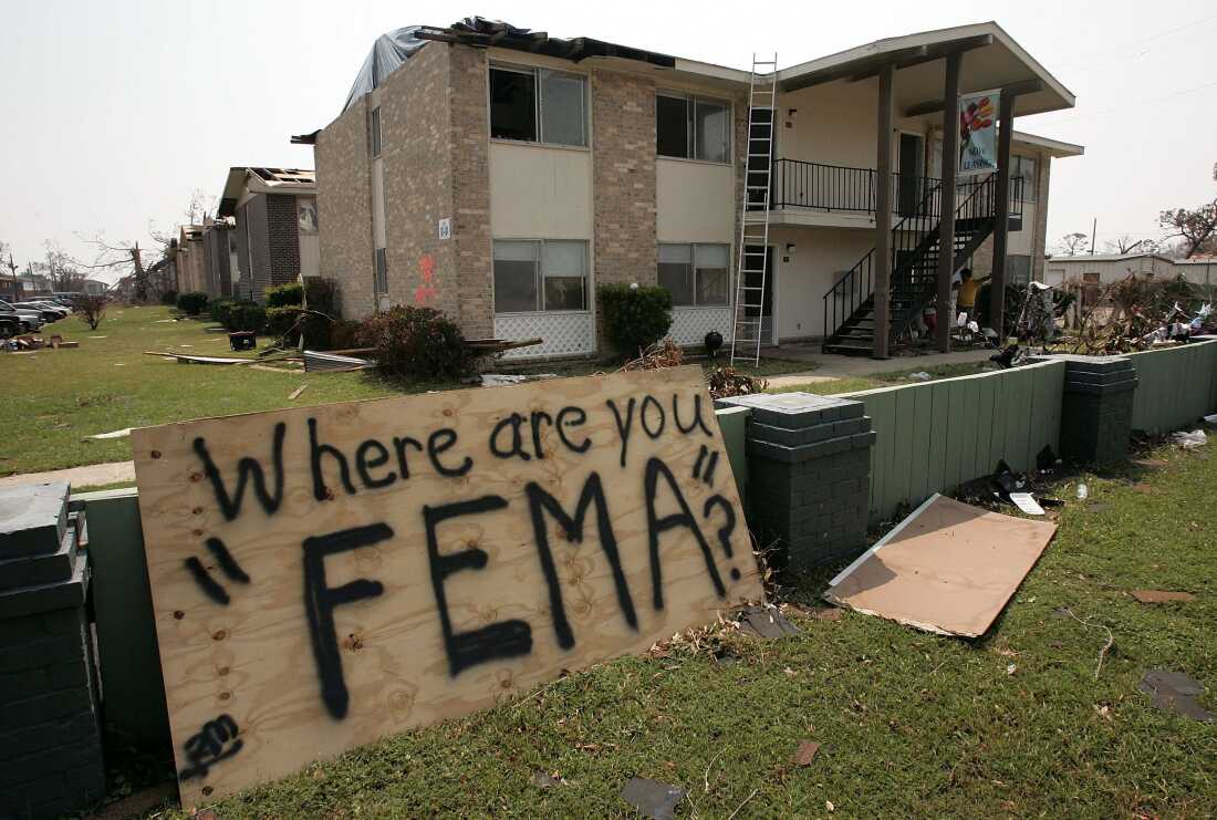 A sheet of plywood with the words "Where are you FEMA?" spray-painted on it leans against a wall in front of an apartment complex with hurricane debris all around. 