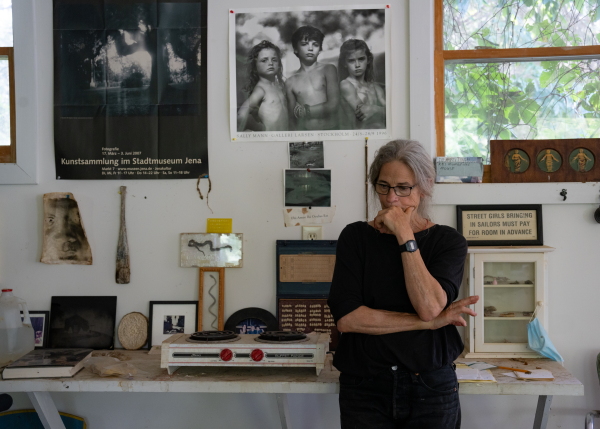 Sally Mann is shown standing in her collodion studio beneath a print of the 1989 photograph titled "Emmett, Jessie and Virginia."