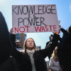 A demonstrator at a "Stand Up For Science" rally holds a homemade poster in the air that reads "knowledge is power not waste" in all-caps. Other protesters surround this central protestor.