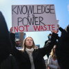 A demonstrator at a "Stand Up For Science" rally holds a homemade poster in the air that reads "knowledge is power not waste" in all-caps. Other protesters surround this central protestor.