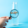 Close-up photograph of a hand holding magnifying glass over a bottle of water. Microplastic particles seen floating in glass bottle with water.