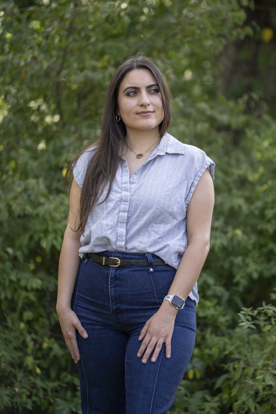 Nusheen Farahani is standing outdoors, with green foliage in the background. She is wearing blue pants and a light-blue sleeveless shirt.