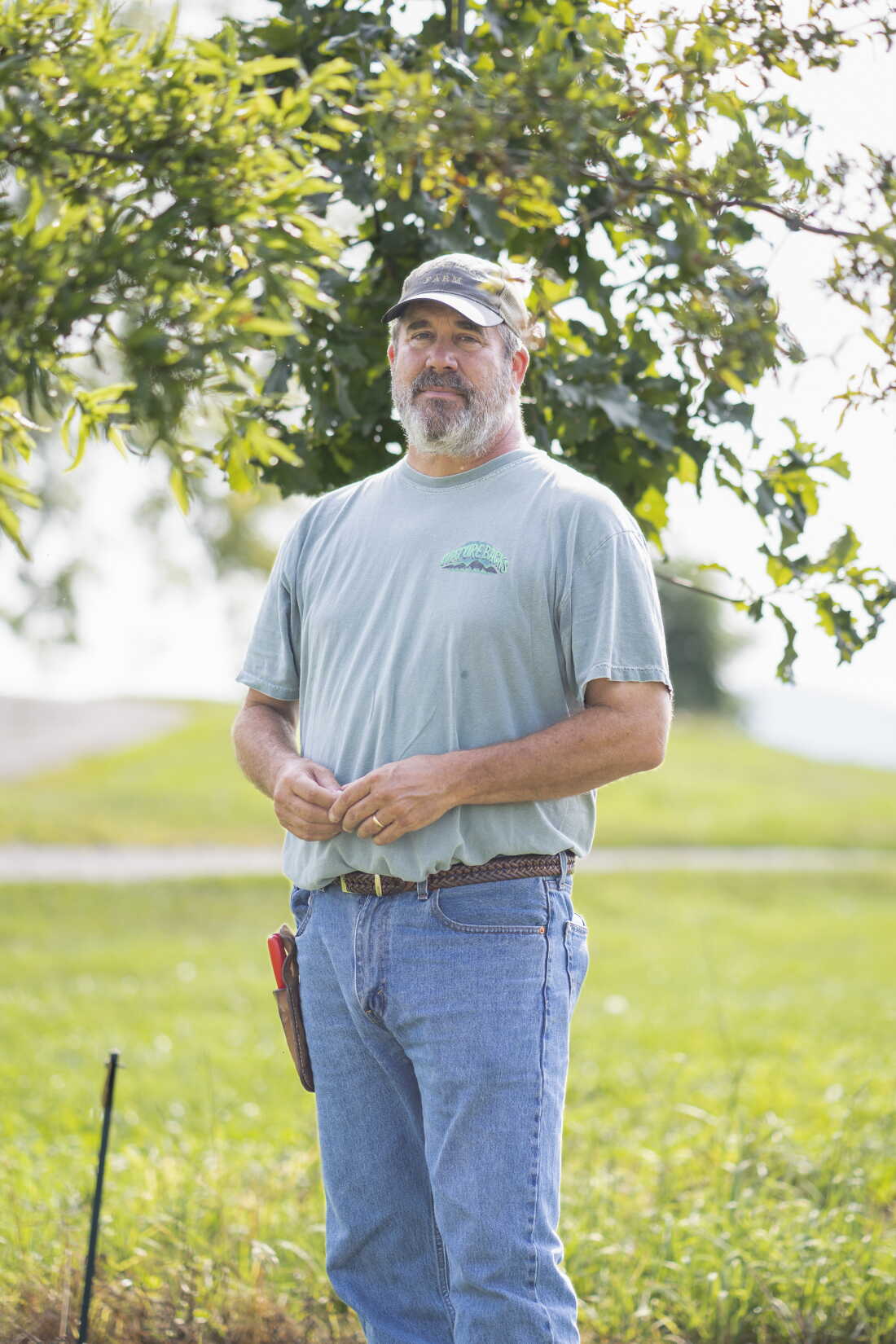 Steve Black stands outdoors. He's wearing blue jeans, a T-shirt and a baseball cap. Green grass is in the background, and tree branches with green leaves hang in the background.