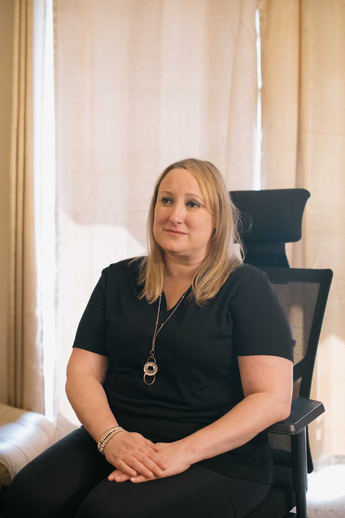 ALT TEXT: A portrait of a 43 year old white woman with shoulder length blonde hair. She sits in a chair with her hands placed on her lap. She is wearing all black and has a neutral expression.