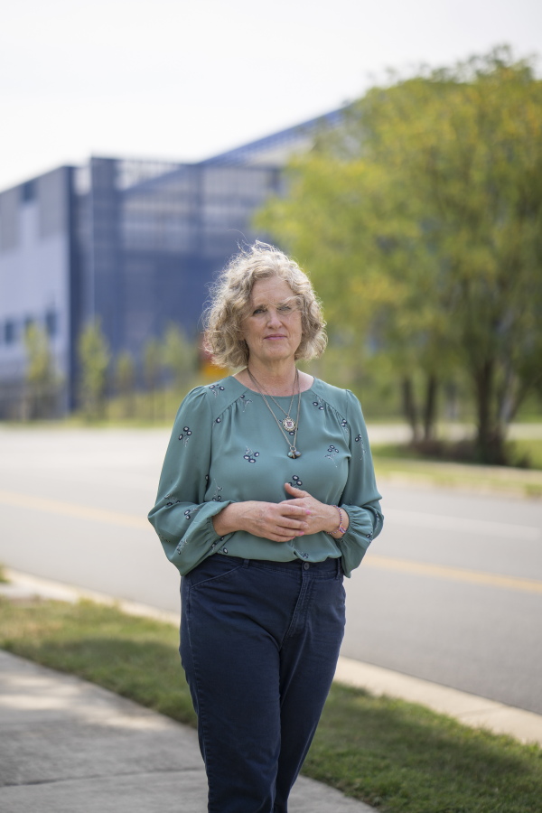 Juli Briskman stands on the sidewalk along the street in front of the Vantage VA2 data center in Sterling. She's wearing blue pants and a teal long-sleeved shirt. The data center is a large blue building in the background.
