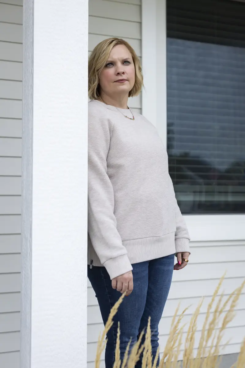 Samanthya Marlatt, who has cerebral palsy and spent time in foster care, lives in Iowa. She has chin-length blonde wavy hair and is wearing a cream colored sweater while standing outside her home.