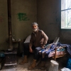 Joseph Elias Issa, 56, sits on a day bed next to a wood burning stove in a small cement building. He has a white beard and a scarf wrapped on his head.