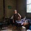 Joseph Elias Issa, 56, sits on a day bed next to a wood burning stove in a small cement building. He has a white beard and a scarf wrapped on his head.