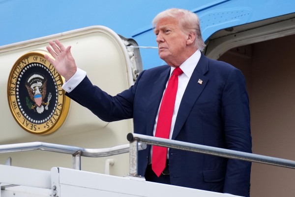 President Trump waves as he emerges from the airplane's door and stands at the top of the steps that lead up to the airplane's door.