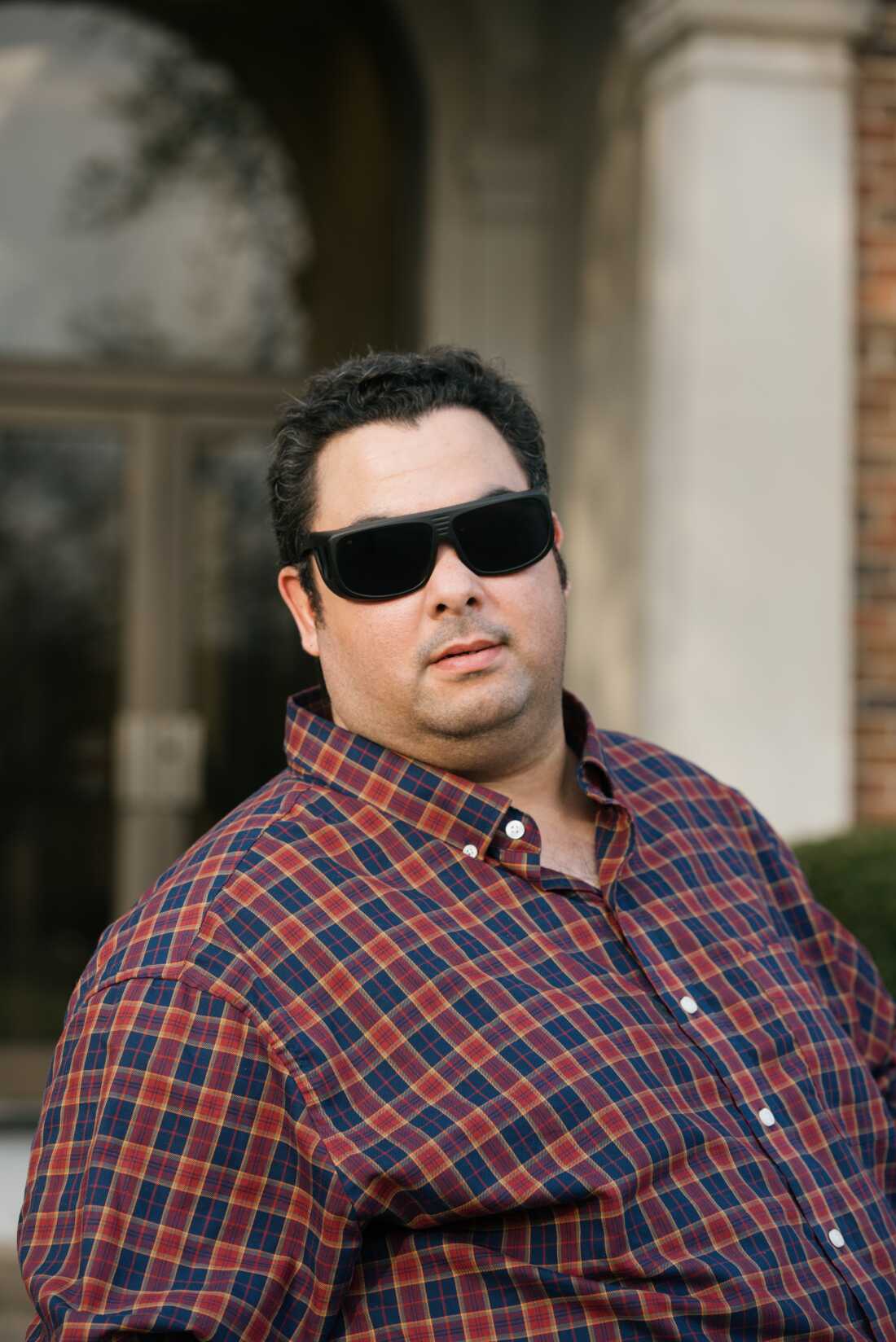 ALT TEXT: A portrait of a 34 year old white man with short, black curly hair. He is wearing dark glasses and a maroon and blue plaid, buttoned shirt. 