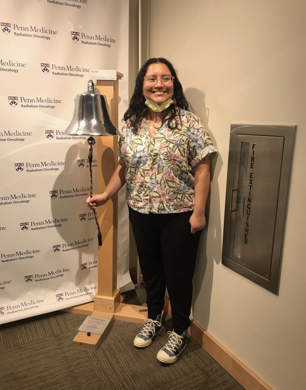 Lourdes Monje rings a big silver bell standing in front of wallpaper that says "Penn Medicine."