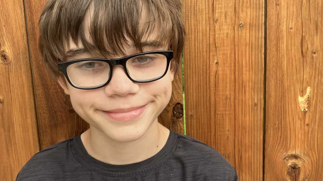 This photo shows sixth-grader Levi Chowske. He's wearing a black T-shirt and black-framed glasses. He's standing against a brown wooden fence.