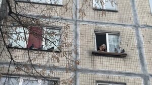 Nelia Stepanivna Thomashevska, 80, waves from her kitchen window, which is on the fourth floor of an apartment building. Two birds are perched on the windowsill.