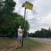 A man uses a long metal pole with a bright yellow sign taped to the end to block the view of a camera mounted on a pole next to a road. The sign says "Deflock Crystal dot com."