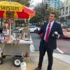 A man wearing a suit and a red tie stands on a city street next to a hot dog cart with a colorful red and yellow umbrella.