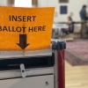 A ballot box has a sign that says "insert ballot here" with an arrow pointing to the slot.