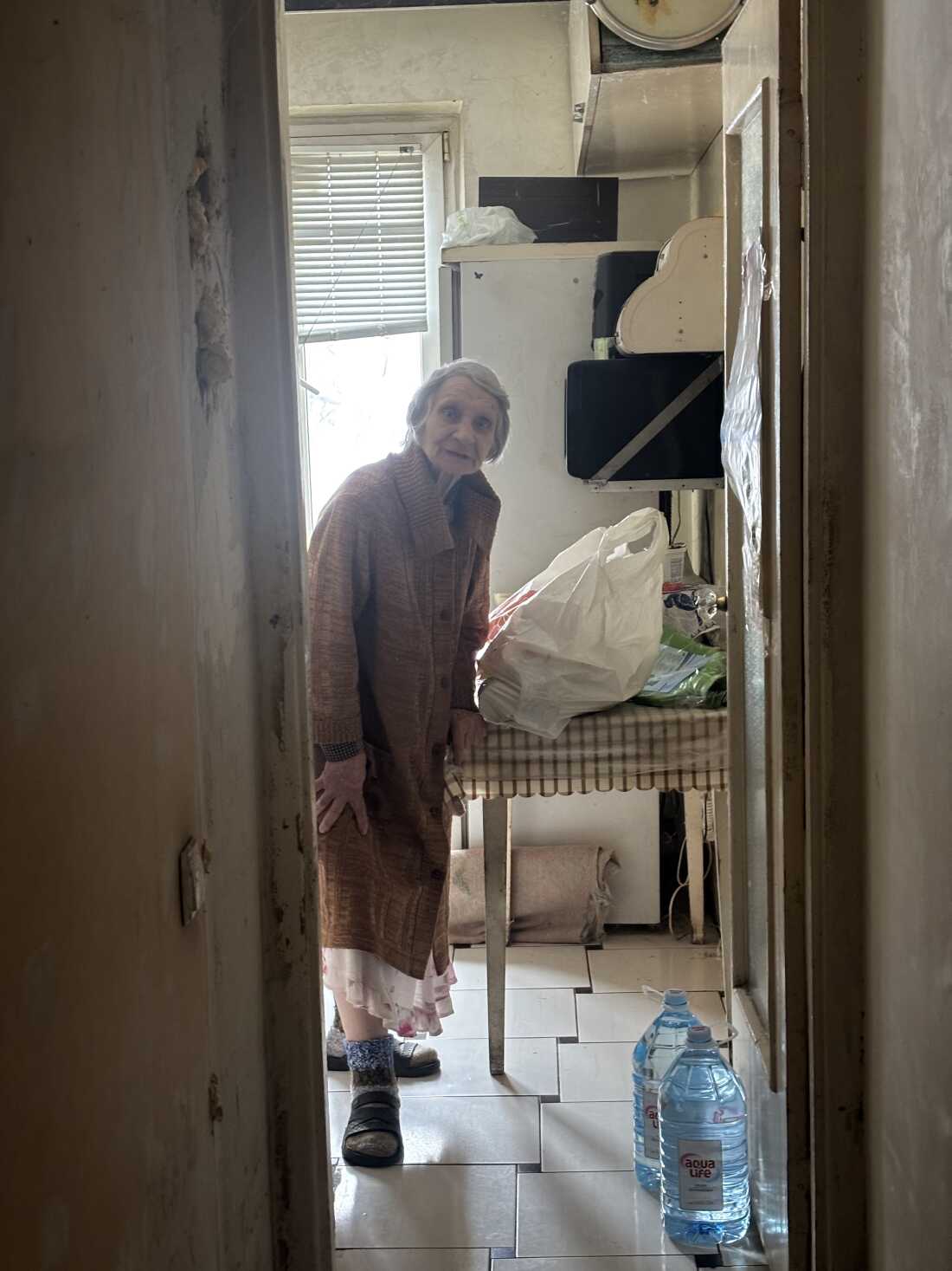  Photographed through a doorway within her home, Nelia Stepanivna Thomashevska, 80, is wearing a long brown sweater coat and is standing next to a table with a large plastic bag on it. Two large bottles of water are on the floor.