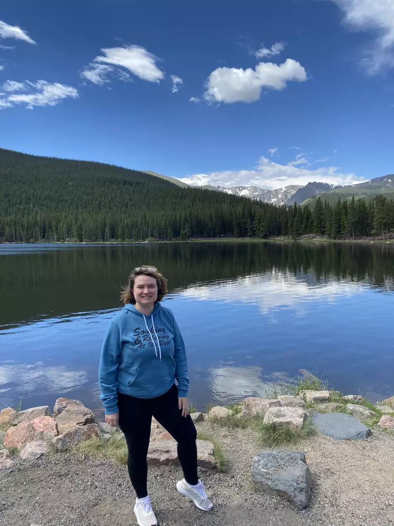 Molly Quinn stands at the shore of a beautiful lake. Pine trees and mountains are in the background.