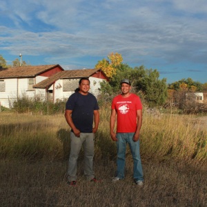 Lonny and Teyon Fritzler stand outside their childhood home. Plywood covers the windows of the white house, and tall gold-colored grass is high in the yard between the brothers and the house.