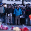 Community members stand shoulder to shoulder, some holding hands, at a vigil. There are bouquets of flowers on the ground.