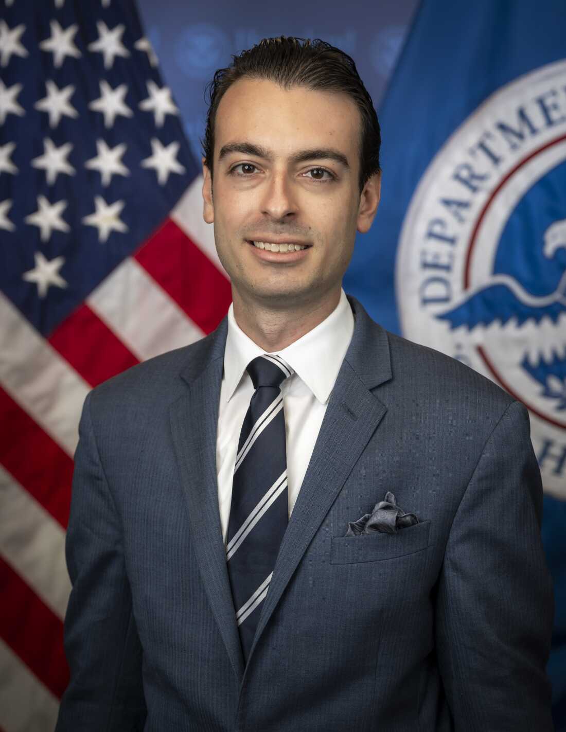 This photograph shows Paul Ingrassia photographed from astir the waist up. He's wearing a suit and tie, and down him are an American emblem and a Department of Homeland Security flag.