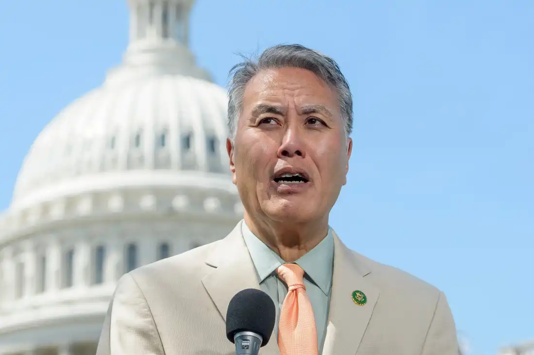 With a microphone in front of him, Representative Mark Takano speaks outdoors on Capitol Hill in 2025. He's wearing a beige suit jacket, and the Capitol dome rises behind him against a light blue sky.