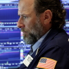 A trader works on the floor of a stock exchange. He is wearing a navy blue blazer, and screens displaying stock market information are behind him.